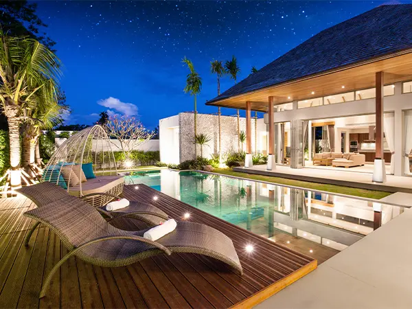 An expensive home with a pool deck with lighting and a couple of long chairs