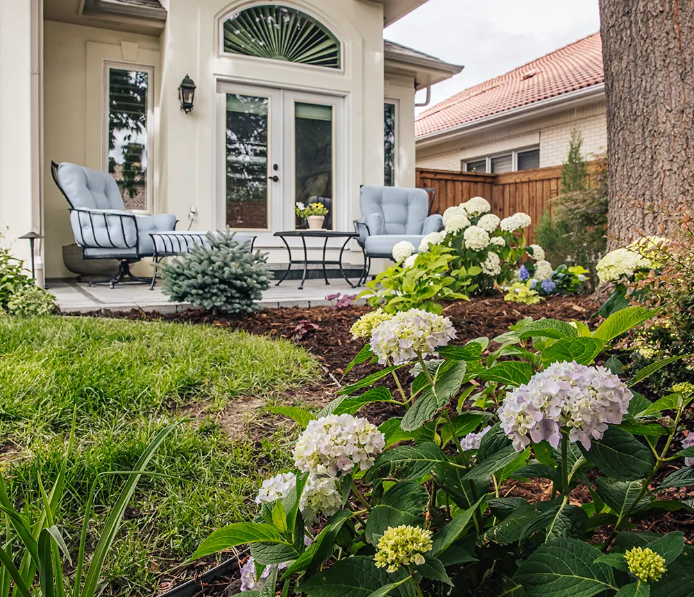 Closeup of home landscape with flowers, a big tree, and a custom paver patio with comfortable chairs