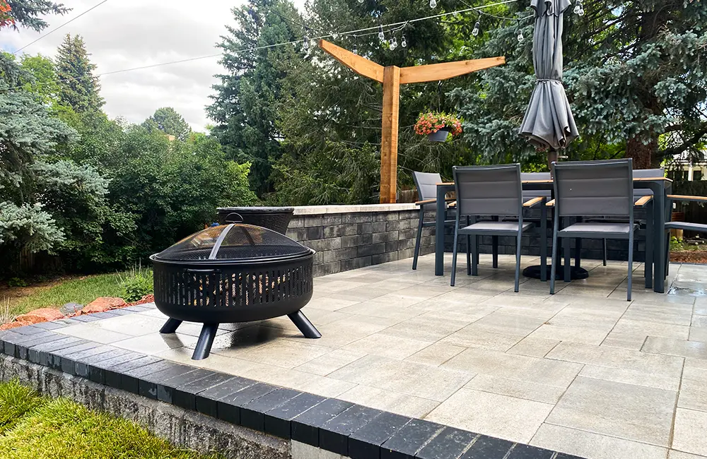 Big custom paver patio with firepit, table with chairs, and elegant black retaining wall