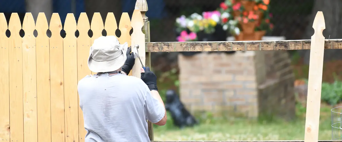 A man in a hat and gloves installing a wooden picket fence in Arvada, in a backyard garden, with colorful flowers in the background.