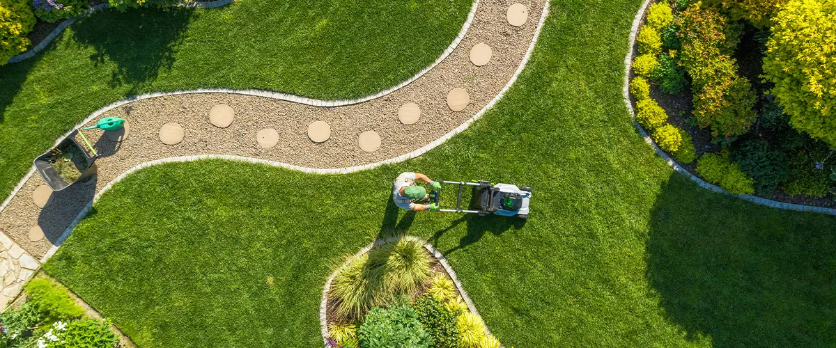 Aerial view of a gardener mowing a well-manicured lawn with winding stone paths and lush landscaping in a beautifully designed garden.