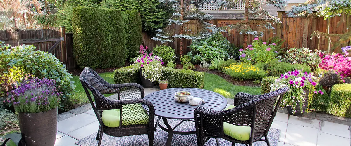 Charming backyard garden with lush greenery, colorful flowers, and cozy outdoor seating area with wicker chairs and a round table.