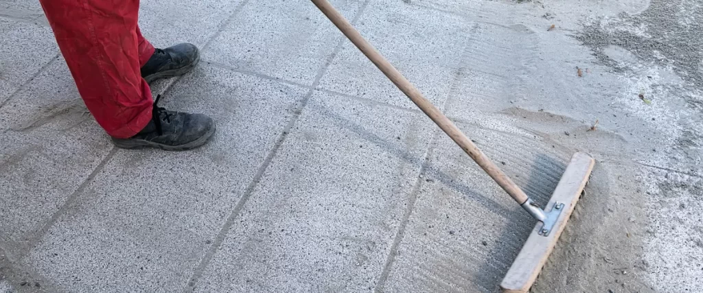 Worker using a large brush to spread sand over a newly installed concrete surface