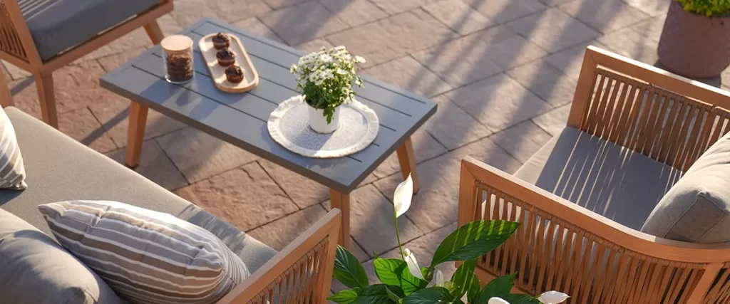 Outdoor patio with wooden lounge chairs, a gray coffee table, and greenery, set on a stone paver surface with soft sunlight.