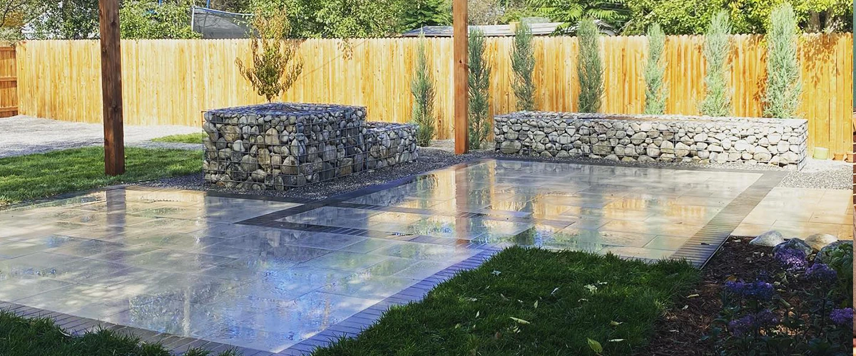 Glossy finish paver patio with gabion retaining walls and pergola posts surrounded by fresh landscaping.
