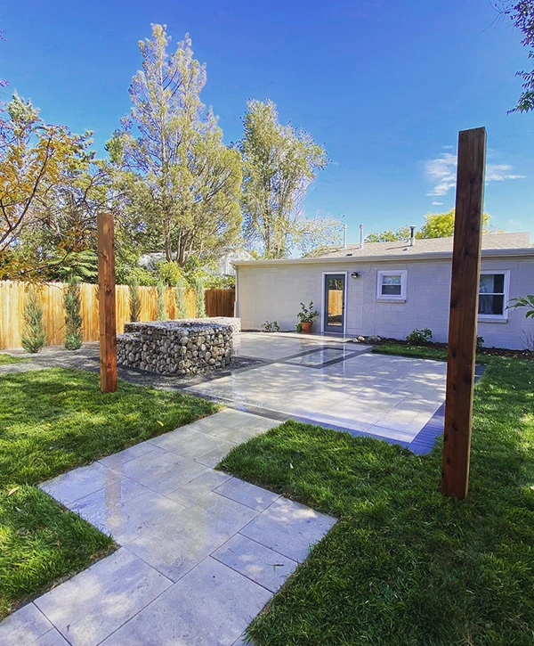 Modern backyard with a paver patio, stone path, and gabion-style retaining wall surrounded by grass.