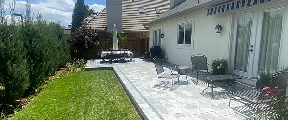 Elegant backyard patio with gray stone pavers, black metal furniture, lush green lawn, and white siding exterior, creating an ideal outdoor living space under a blue sky.