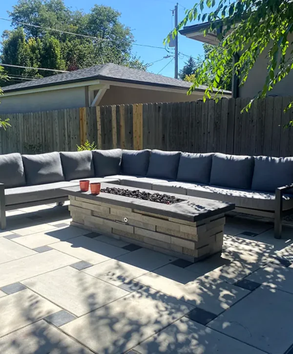 Modern outdoor patio with a gray sectional sofa, stone fire pit, geometric tile flooring, wooden privacy fence, and lush greenery.