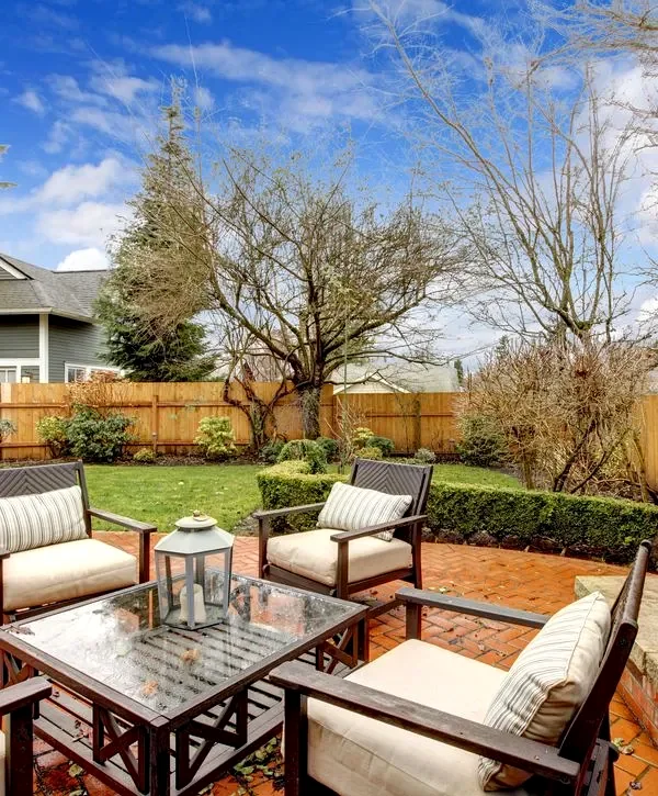 Cozy brick patio featuring teak wood chairs with cream cushions, a glass-top table with a decorative lantern centerpiece, surrounded by a wooden fence and manicured lawn.