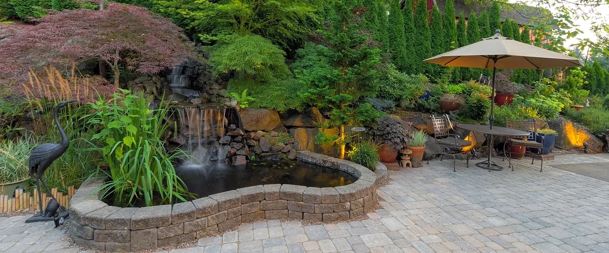 Elegant backyard patio with paver stone flooring, a small koi pond with a waterfall built into a stone retaining wall, and a shaded outdoor dining set under a beige patio umbrella.