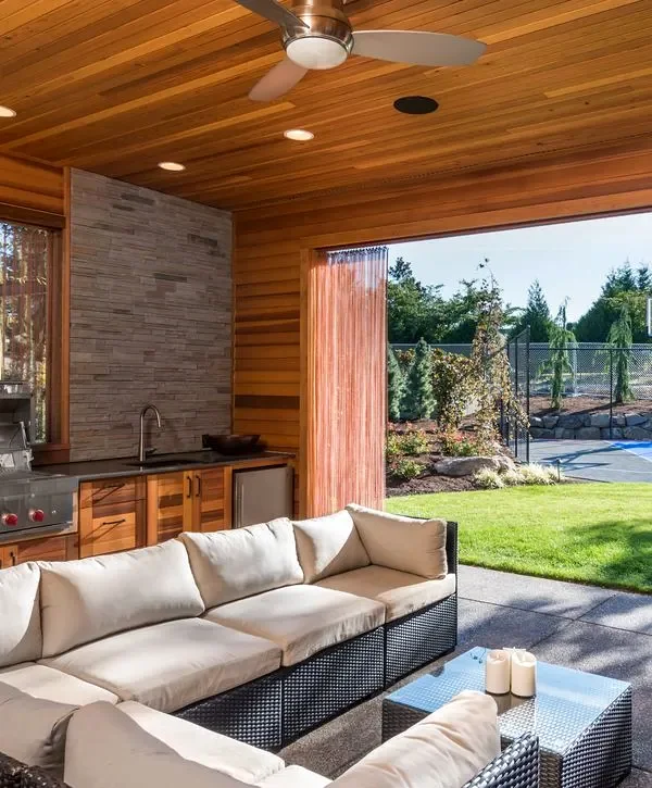 Luxurious covered patio featuring a cedar wood ceiling with a fan, a black wicker sectional sofa with cream cushions, and a stainless steel outdoor kitchen with a stone backsplash.