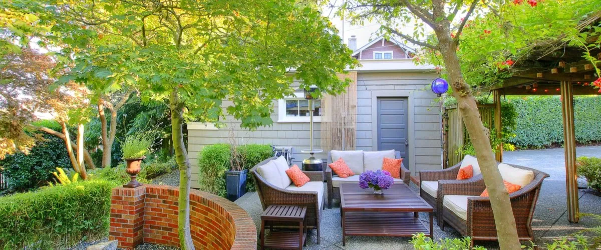 Shaded backyard patio with comfortable wicker furniture set, cream cushions, and orange pillows, situated under a pergola and surrounded by lush trees and greenery next to a modern gray shed.