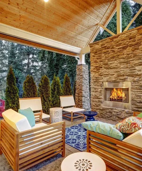 Cozy covered patio with a stacked stone fireplace, modern slatted wooden outdoor furniture, a blue patterned rug, a cedar wood ceiling, and tall greenery in the background.