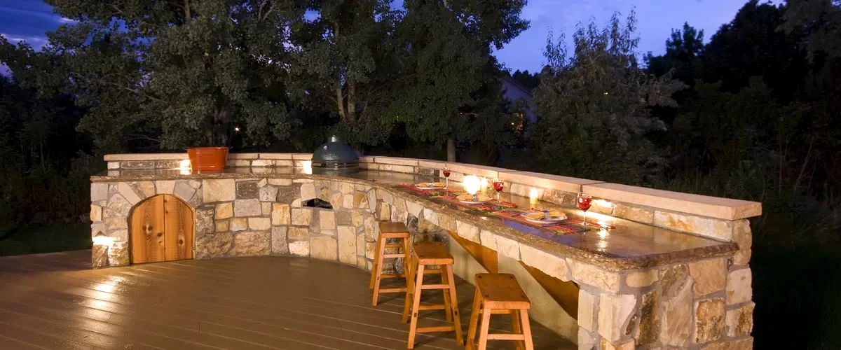 Rustic stone outdoor kitchen with a curved countertop, integrated grill, and wooden door, featuring three wooden bar stools and ambient lighting on a deck at twilight.