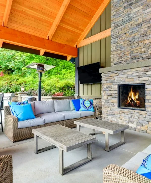 Luxury covered patio with a stacked stone fireplace, modern wicker furniture with bright blue throw pillows, an outdoor heater, and a wooden vaulted ceiling, perfect for year-round entertaining.