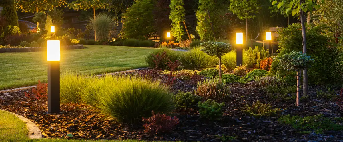 Sleek black bollard-style landscape lighting fixtures illuminating a manicured garden border and mulch bed at dusk.