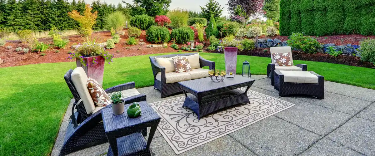 A spacious stone patio featuring dark wicker sofas, floral pillows, and a decorative rug, showcasing a modern backyard landscape design.