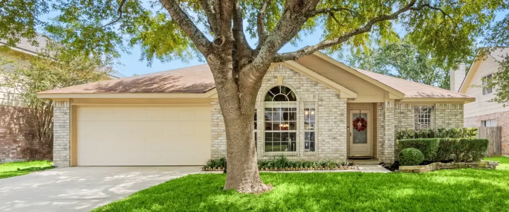 a single-story residential home with a beige brick exterior, a light-colored garage door, and a mature tree in the front yard