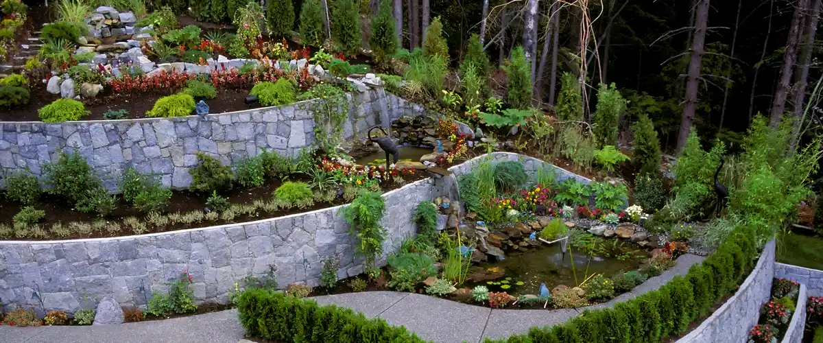 Large-scale multi-tiered grey stone retaining walls on a hillside with a garden pond, water fountain, and decorative crane statues in a professional hardscape.