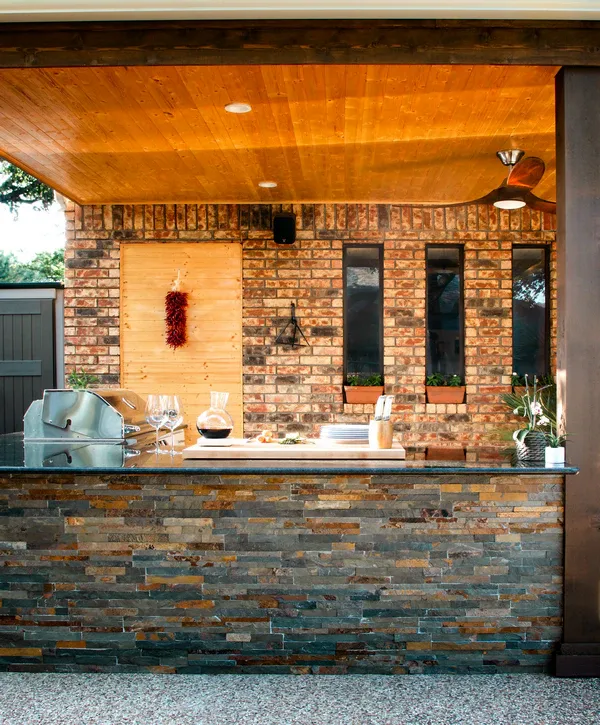 Luxury outdoor kitchens in Wheat Ridge, CO showcasing a dark stone veneer island with a built-in grill, black countertops, wine glasses, and a cutting board beneath a natural wood ceiling.
