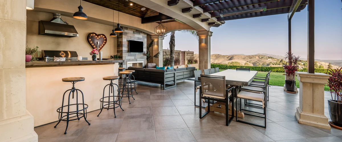 A spacious, luxurious covered patio featuring a fully equipped outdoor kitchen with barstool seating, alongside an elegant dining table and a comfortable lounge area with a TV, overlooking scenic rolling hills.