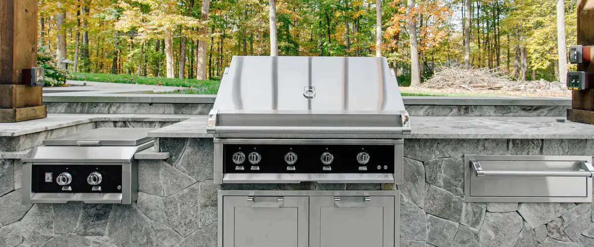 A light grey stone outdoor kitchen featuring a large built-in stainless steel grill with black control panels, a side burner, and countertops set against a wooded autumn background.